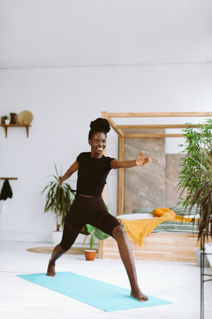 Vertical smiling interracial woman do yoga on mat at home. Keep hands straight above head in Virabhadrasana Warrior twoの写真素材