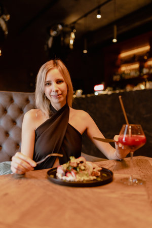 Beautiful blonde in evening dress enjoys signature salad and cocktail in night restaurant. Have great evening.の写真素材