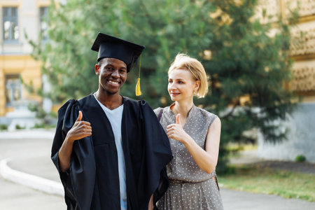 Portrait of afro american student in graduation mantle and hat standing outdoors with happy middle aged woman. Higher education, graduate from university, like, finger up, ceremonial celebrationの写真素材