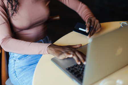 Unrecognizable African-American woman sitting at table, working doing online shopping on laptop, using credit card.の写真素材