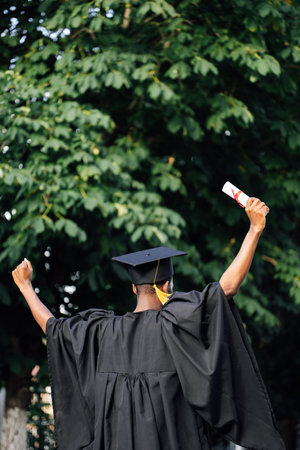 Afro american graduate from university in black mantle and hat stand with back outdoors among trees and holding higher education diploma with raised arms. Start in life, graduation, freedomの写真素材