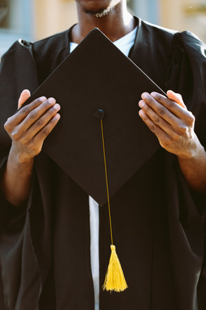 Hands of african american student in black mantle holding graduation hat closeup. Young specialist, graduate from university, higher education, start in life, ceremonial celebrationの写真素材