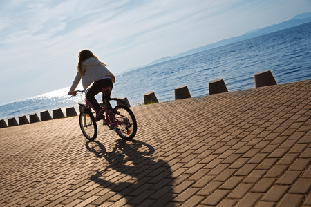 Girl rides a bike on the promenade by the sea at sunset. Focus on the pavement. On image superimposed filter Color Lookup.の写真素材
