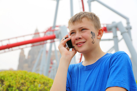 Cute boy talking on a cell phone at an amusement park. Drawing a dragon on her cheek.の写真素材