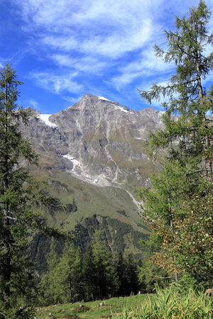 Forests, mountains and rocky peaks of the Alps below. Remains of unmelted snow in mountains. Rock fragments on a green field.の写真素材