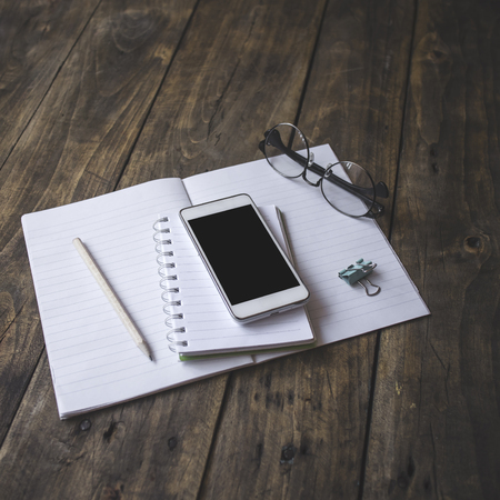 Home office tabletop: papers and notebooks, reading glasses, smart phone, pen on an old wooden board background. Vintage lifestyle.の写真素材