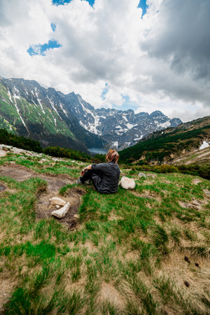 A tourist girl is relaxing in the mountains, sitting high on the mountain with her back to us and looking at the horizonの写真素材