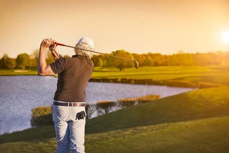 Senior golf player shooting ball at sunset, with lake in background.の写真素材
