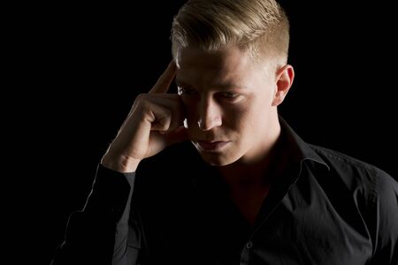 Low-key portrait of young handsome man with hand at temple looking aside, isolated on black background.の写真素材