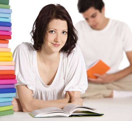 Pretty young woman lying on floor for reading and looking straight with stack of colorful books next to her and young man in background reading, isolated on white background.の写真素材