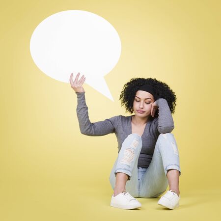 Unhappy woman being sad about being lonely holding a white blank paper speech bubble with copy space for text, isolated on yellow background.の写真素材