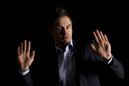 Low-key portrait of young clueless business person in dark suit sitting at office desk being confused, isolated on black background with copy-space.の写真素材