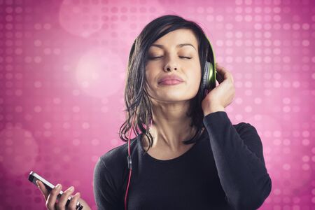 Young joyous girl listening to music with headphones and mp3 player with eyes closed, in calm and relaxed mood, isolated on pink disco background.の写真素材
