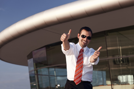 Happy businessman showing both thumbs up, office building in background.の写真素材