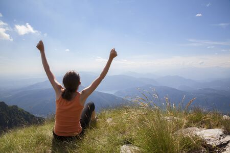 Happy woman with raised arms sitting on mountain top looking at mountain landscape and blue sky on Monte Generoso.の写真素材