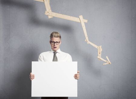 Concept: Business crisis. Worried businessman holding white empty signboard with space for text in front of business graph with negative trend, isolated on grey background.の写真素材