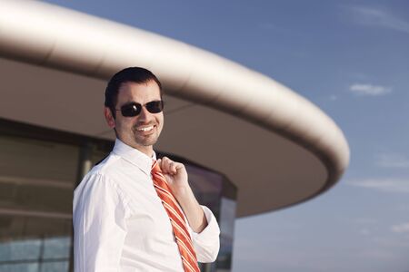 Portrait of smiling young business person in white shirt, orange tie and sunglasses standing in front of office building.の写真素材