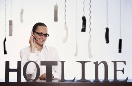 Smiling female phone operator in call centre sitting at desk and talking with headset providing customer support with many phone receivers hanging in background.の写真素材
