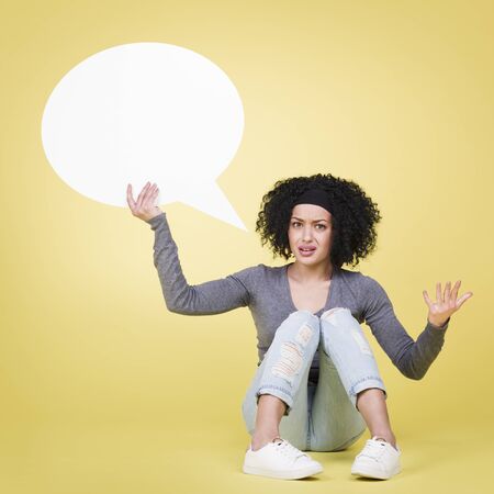 Wondering woman being irritated holding a blank white paper speech bubble with empty copy space, isolated on yellow background.の写真素材