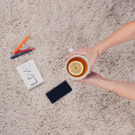 A woman holds hands a cup of tea with lemon, phone, pen and notebook lying on the carpet. Top viewの写真素材
