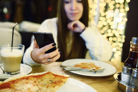 Woman typing write message on smart phone in a modern cafe. Cropped image of young pretty girl sitting at a table with pizza using mobile phone. Tonned. Selective focus.の写真素材