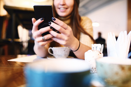 Woman typing text message on smart phone in a cafe. Cropped image of young woman sitting at a table with a coffee using mobile phone. Tonned. Selective focus.の写真素材