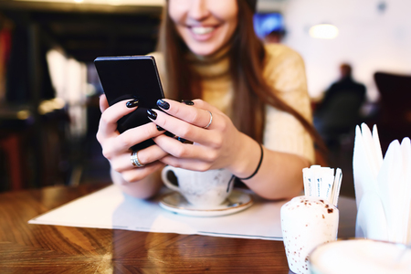 Close up of womens hands holding cell telephone with blank copy space scree for your advertising text message or promotional content, hipster girl watching video on mobile phone during coffee break. Tonned. Selective focus.の写真素材