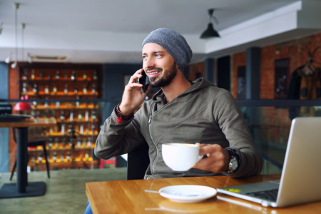 Young handsome hipster man with beard sitting in cafe talking mobile phone, holding cup of coffee and smiling. Laptop on wooden table.の写真素材