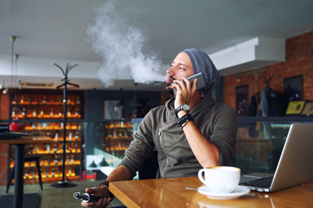 Young handsome hipster man with beard sitting in cafe with a cup of coffee, vaping and releases a cloud of vapor. Talking mobile phone and having a little break.の写真素材
