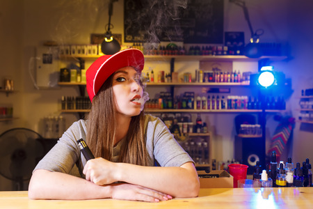 Young pretty woman in red cap smoke an electronic cigarette at the vape shopの写真素材