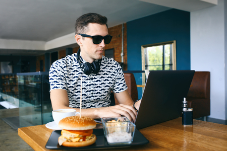 Young handsome hipster man in sunglasses is using laptop in cafeteria. Cup of coffee, electronic cigarette and cheeseburger on wooden table.の写真素材