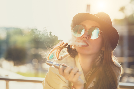 Pretty young hipster woman in black hat vape ecig, vaping device at the sunset. Toned image.の写真素材