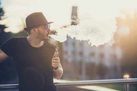 Modern happy young man in hat with a beard fun Vaporizers, In the background, the evening sunset over the city. toned image.の写真素材