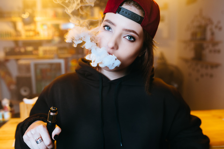 Young pretty woman in red cap smoke an electronic cigarette at the vape shop. Hip-hop style. Closeup.の写真素材