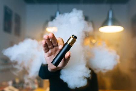 Young beard man show vaping device on his outstretched hand through a cloud of steam. Selective...