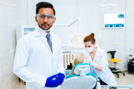 Portrait of a young confident indian male dentist in clinic in front of woman doctor checking teeth to patient. Dental clinic concept.の写真素材