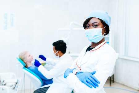 Young African american female dentist in mask in front of dentist checking teeth to patient at clinic. Dental clinic concept.の写真素材