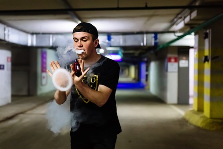 Man in cap smoke an electronic cigarette and releases clouds of vapor performing various kind of vaping tricksの写真素材