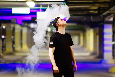 Man in cap smoke an electronic cigarette and releases clouds of vapor performing various kind of vaping tricksの写真素材