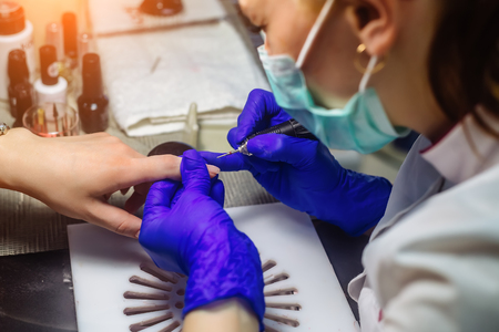 Closeup shot of hardware manicure in a beauty salon. Manicurist is applying electric nail file drill to manicure on female fingers. Selective focus.の写真素材