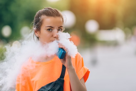 Pretty young hipster woman vape ecig, vaping device at the sunset. Toned image.