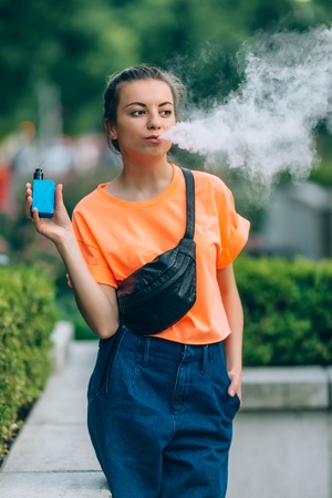 Pretty young hipster woman vape ecig, vaping device at the sunset. Toned image.