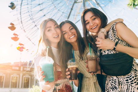 Summer lifestyle portrait multiracial women enjoy nice day, holding glasses of milkshakes. Happy friends inin front of ferris wheel. Best friends girls having fun, joy. Lifestyle. Asian, jewess and caucasian.の写真素材