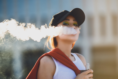Pretty young hipster asian woman in black hat vape ecig, vaping device at the sunset. Toned image. Closeup.の写真素材