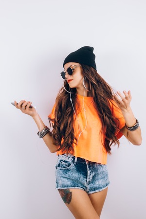 Young hipster beautiful woman in sunglasses wearing in black hat and orange T-shirt listening music in headphones near white wall, holding a cell phone in handの写真素材