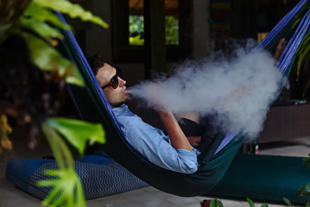 Man resting in a hammock and vaping and letting off steam from an electronic cigaretteの写真素材