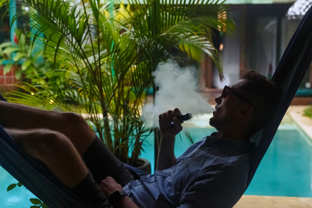 Man resting in a hammock and vaping and letting off steam from an electronic cigarette