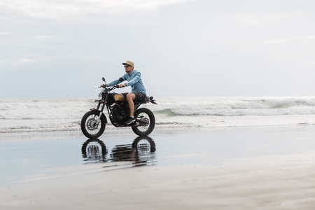 Man in cap riding motorcycle on beach. Moto cross dirtbiker on beach sunset on Bali. Young hipster male enjoying freedom and active lifestyle, having fun on a bikers tour.の写真素材
