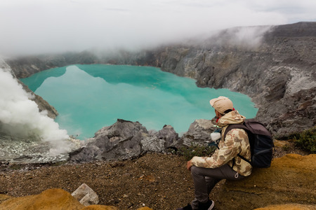 Man tourist looks at the sulphur lake on the Ijen volcano on the island Java in Indonesia. Hiker man with backpack travel on top mountain, travel conceptの写真素材
