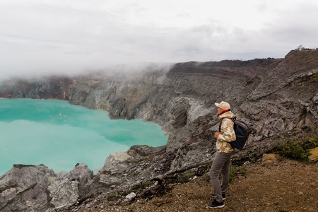 Man tourist looks at the sulphur lake on the Ijen volcano on the island Java in Indonesia. Hiker man with backpack travel on top mountain, travel conceptの写真素材